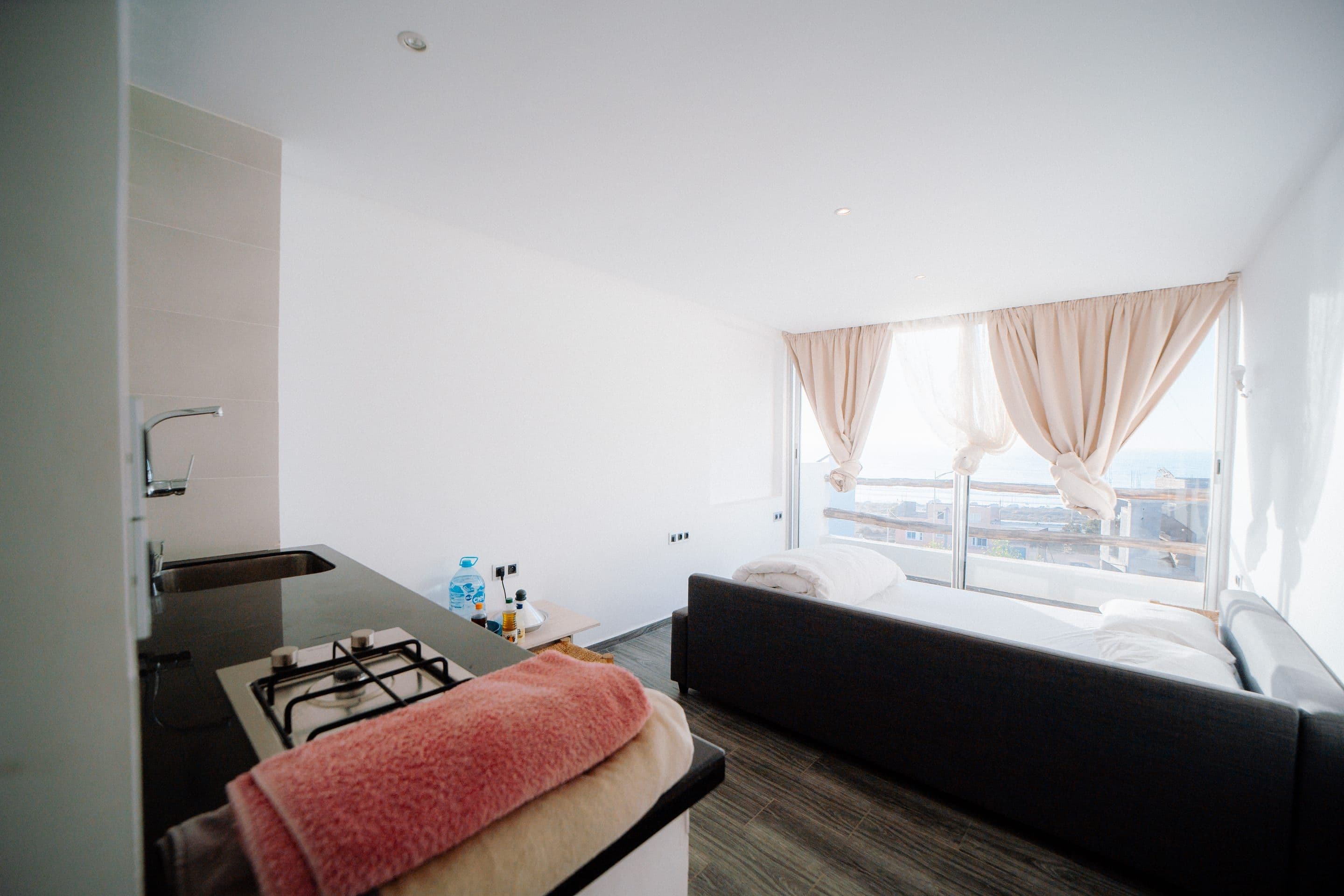 A bright white studio room: bed set beside a floor-to-ceiling window onto the sea, cream curtains drawn back, and a kitchenette with a gas hob along the near wall.
