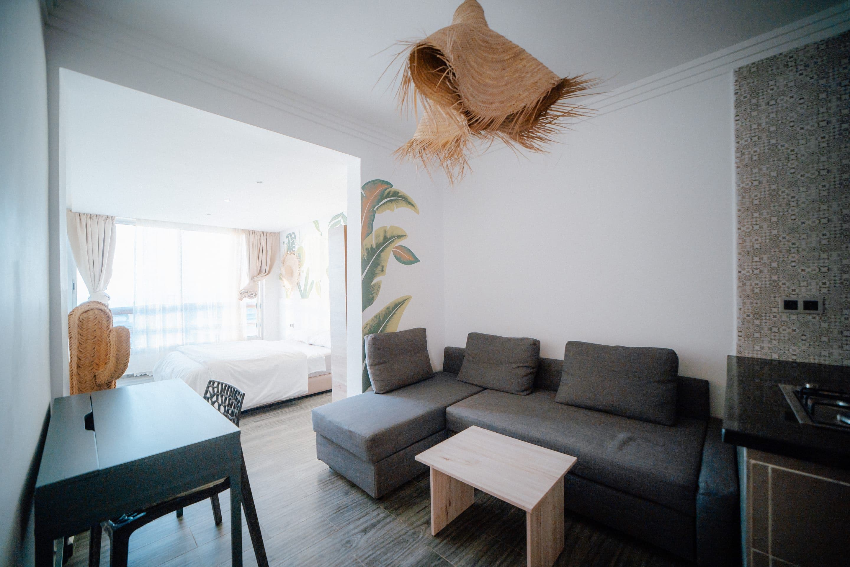 An apartment living room: a grey corner sofa and wooden coffee table, a dining table and chairs, a woven palm pendant lamp, and the bedroom visible through a wide arch.