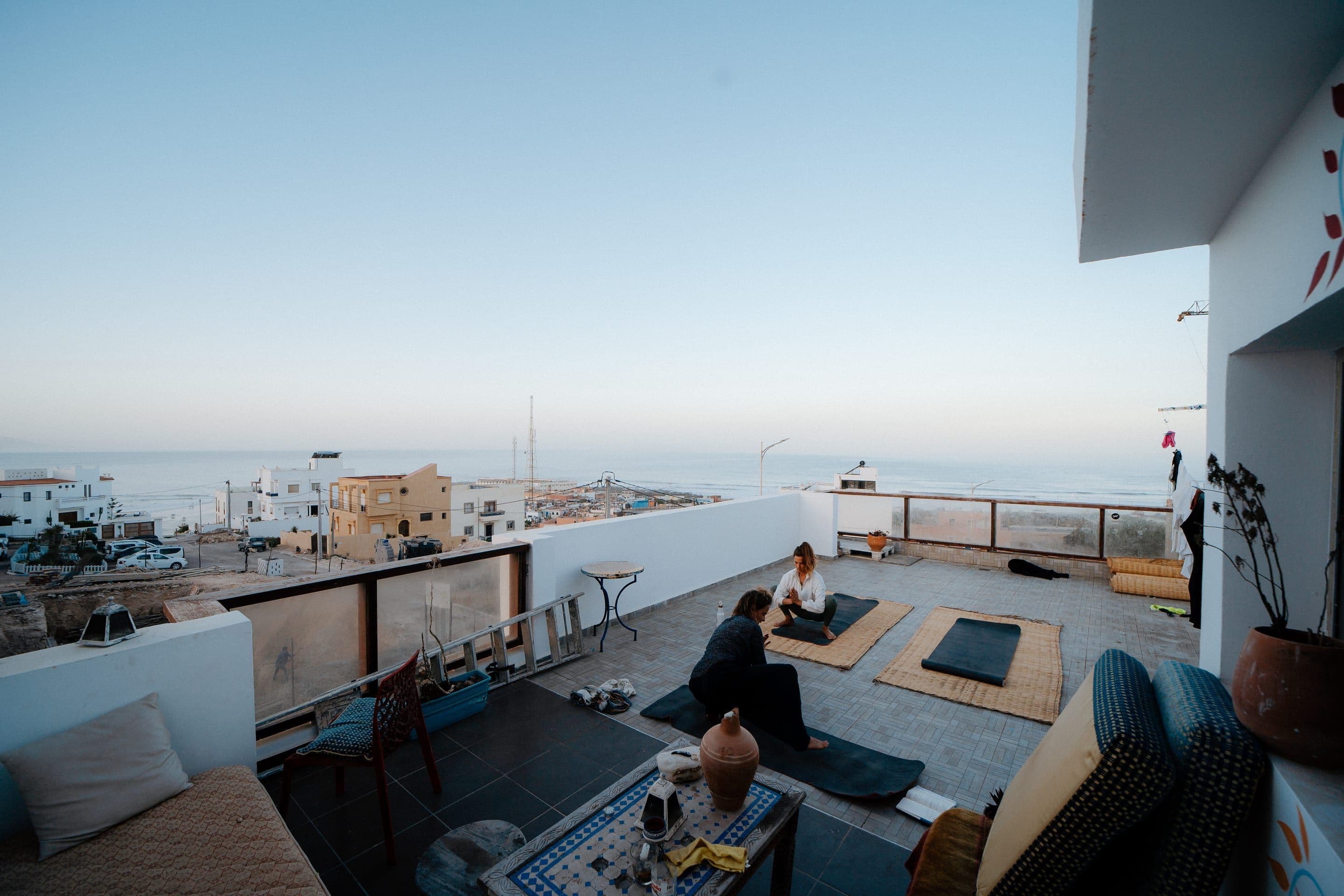 The rooftop terrace at dawn: two people on yoga mats facing the bay, low cushioned benches and a mosaic tea table, with the village rooftops and the ocean beyond.