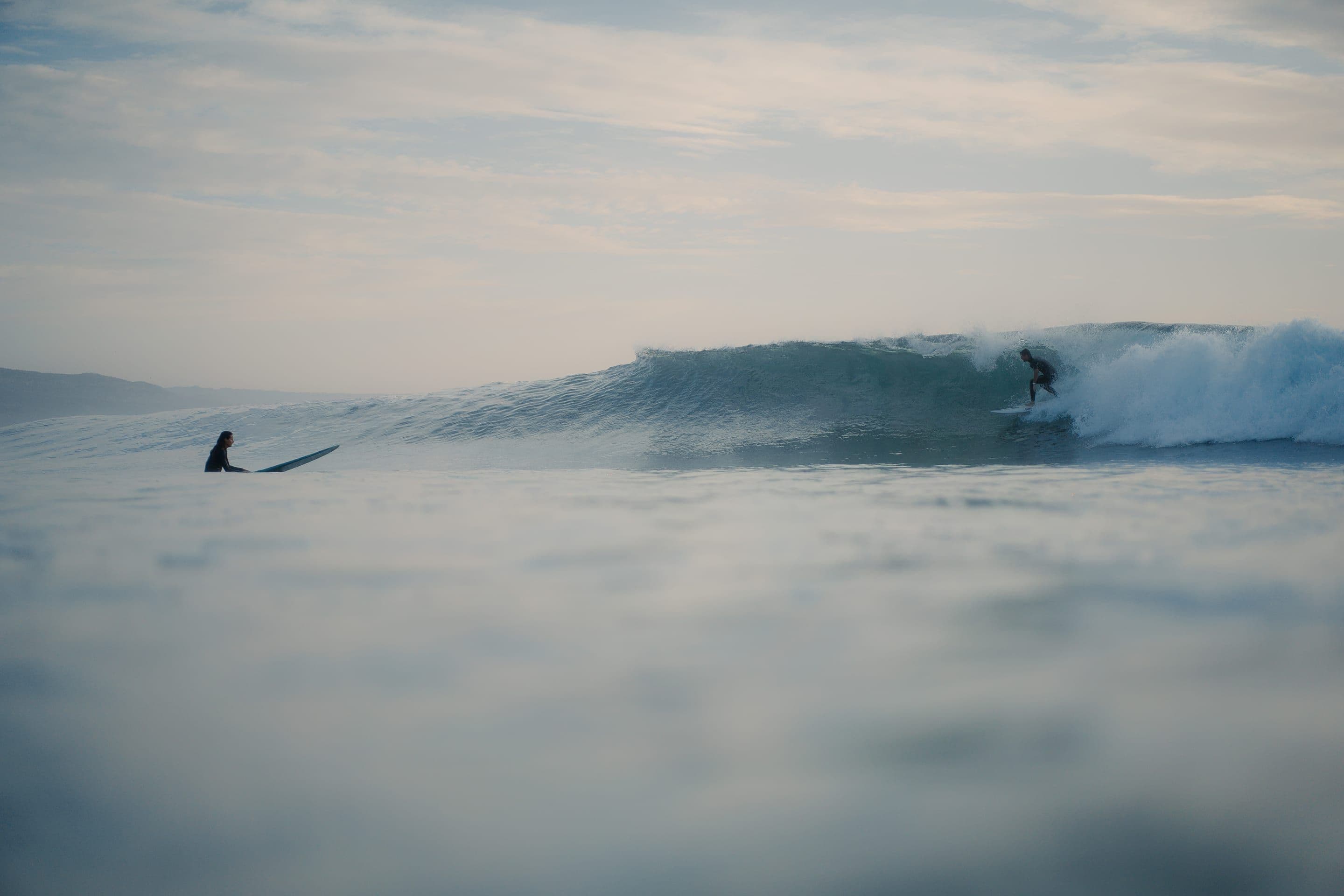 Dawn at Imsouane: a surfer dropping into a clean, glassy right while another waits on a longboard in the flat water inside, the headland soft in the haze behind.