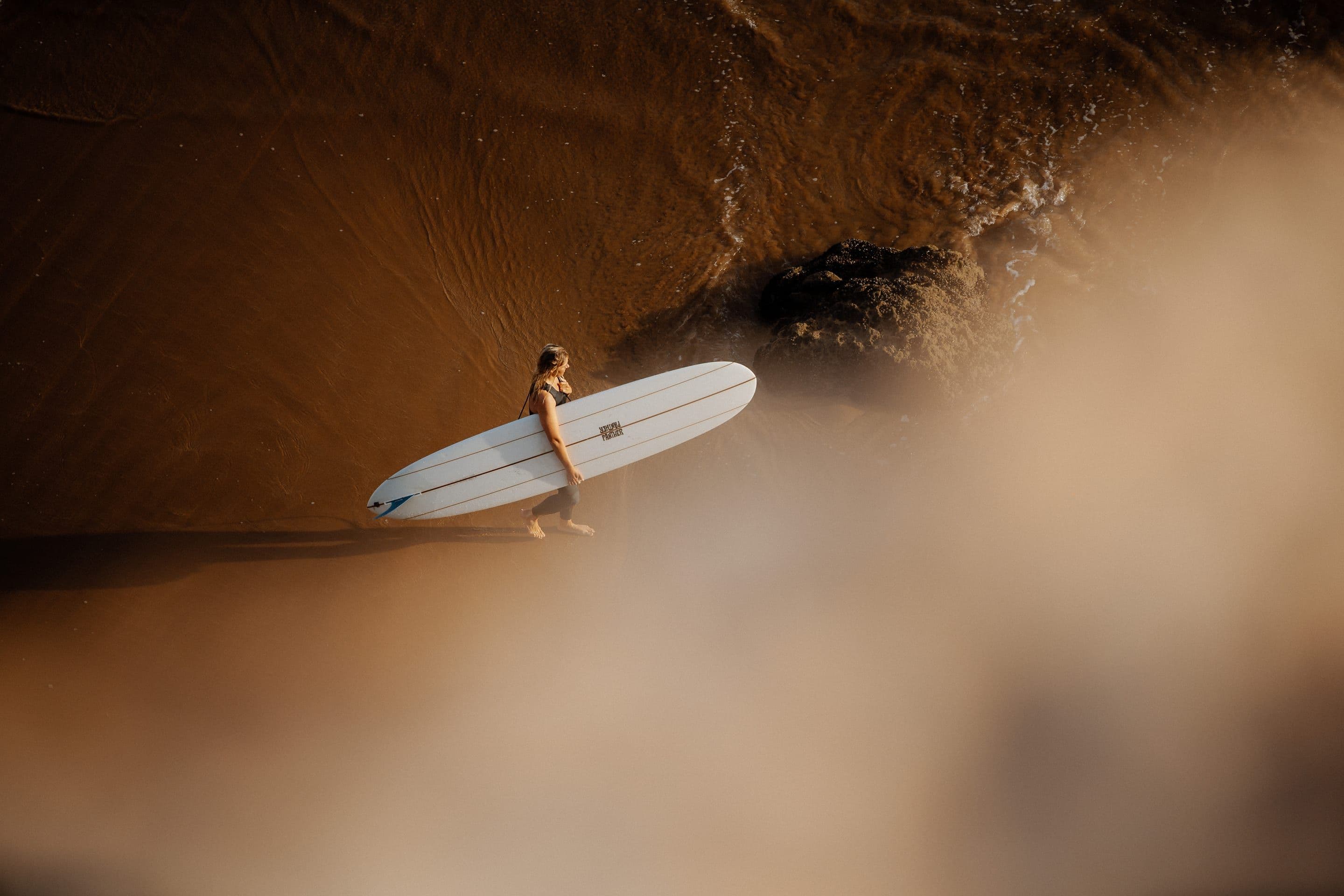 A surfer carrying a white single-fin longboard across wet sand lit gold by the low sun.