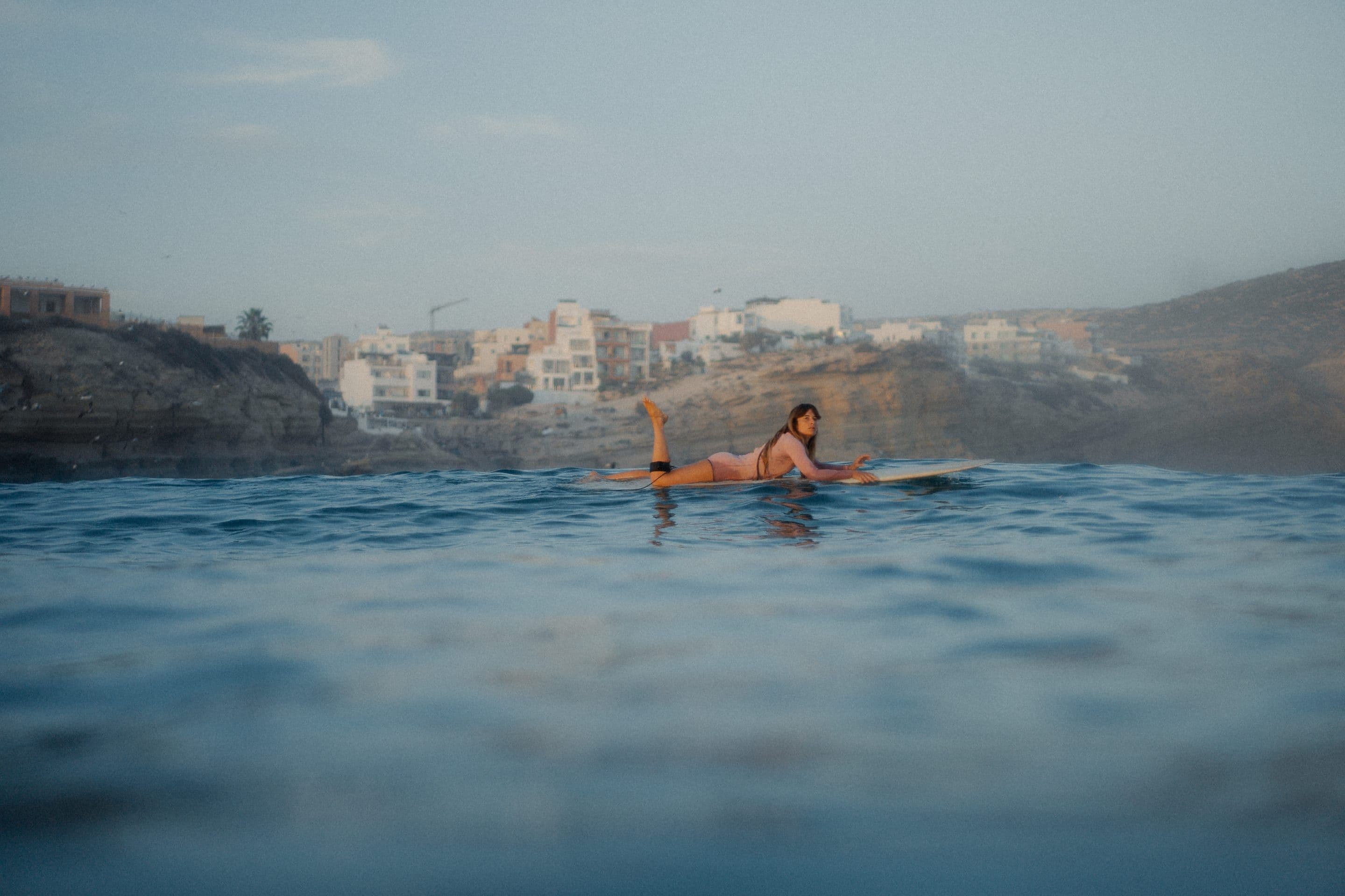A surfer waiting out the back on a longboard in glassy water, looking back over one shoulder, the village stacked along the cliff behind in the last of the light.